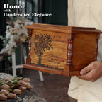 Wooden memorial box with engraved tree design held by a person, with flowers and blurred background.